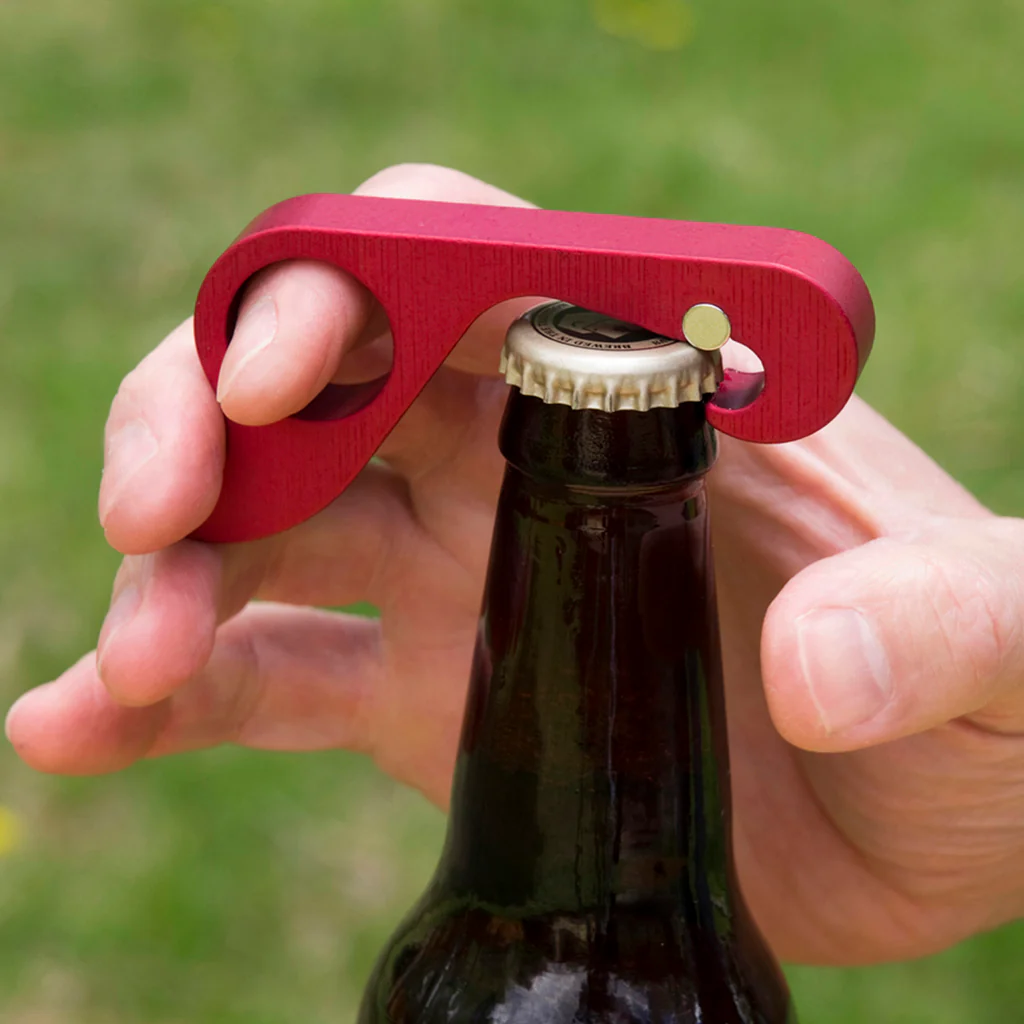 A hand uses a red metal grab opener to gain leverage on a bottle cap.