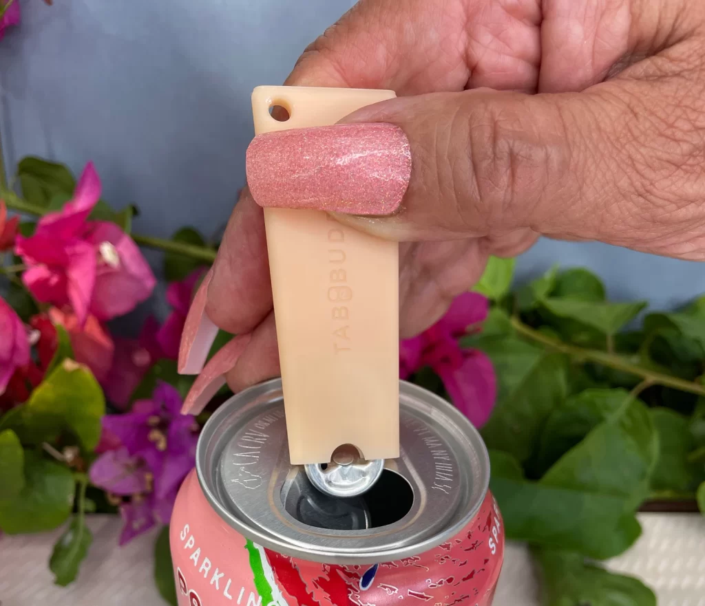 A hand with pink sparkly nails opens a sparkling water can with a salmon colored tab buddy.
