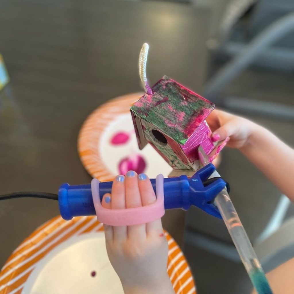 A young girl's hands use a FunctionalHand with a pink EazyHold to hold a paintbrush and paint a small birdhouse.