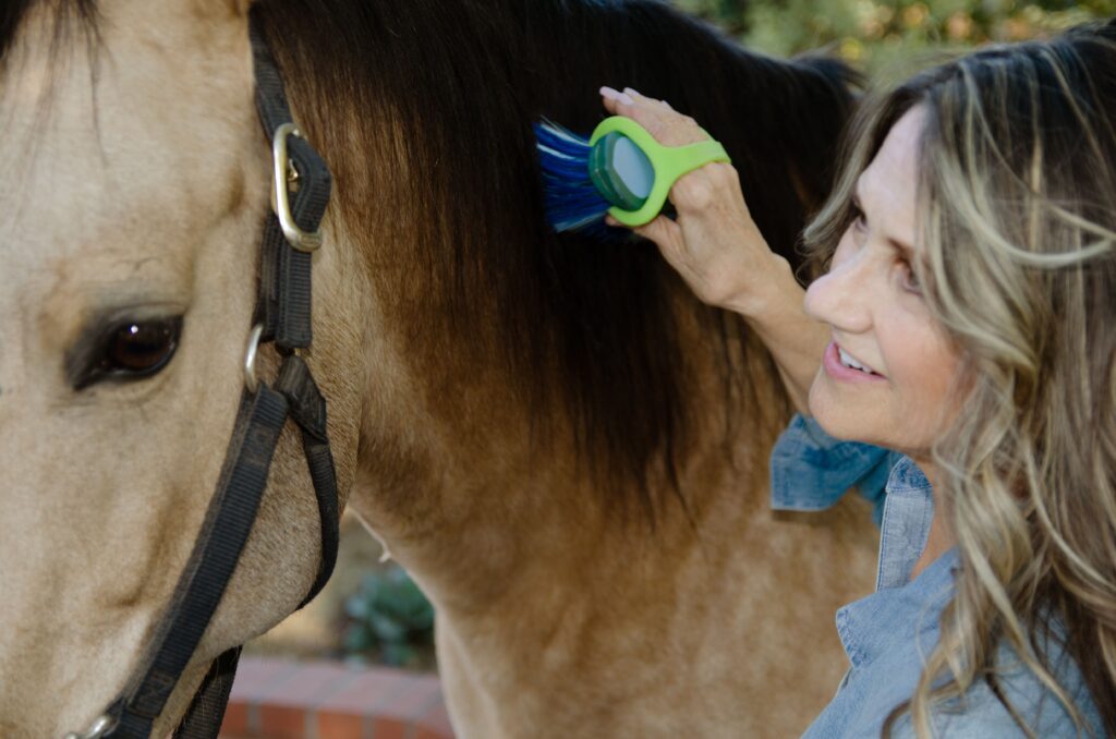 A blonde white woman brushes her horses mane with an EasyHold cuff helping her hold the brush.