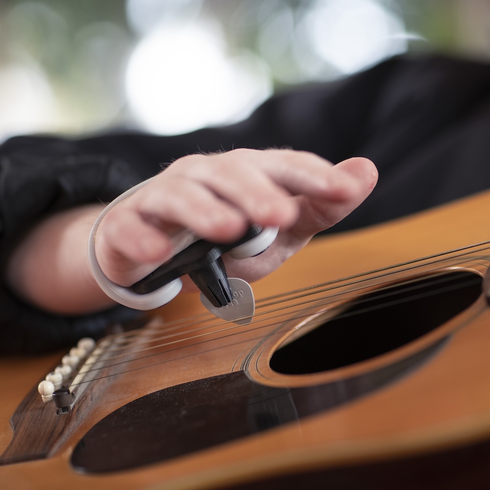 A hand strums a guitar using the Strumling, a pick holding device that uses an EasyHold cuff to attach to the hand.
