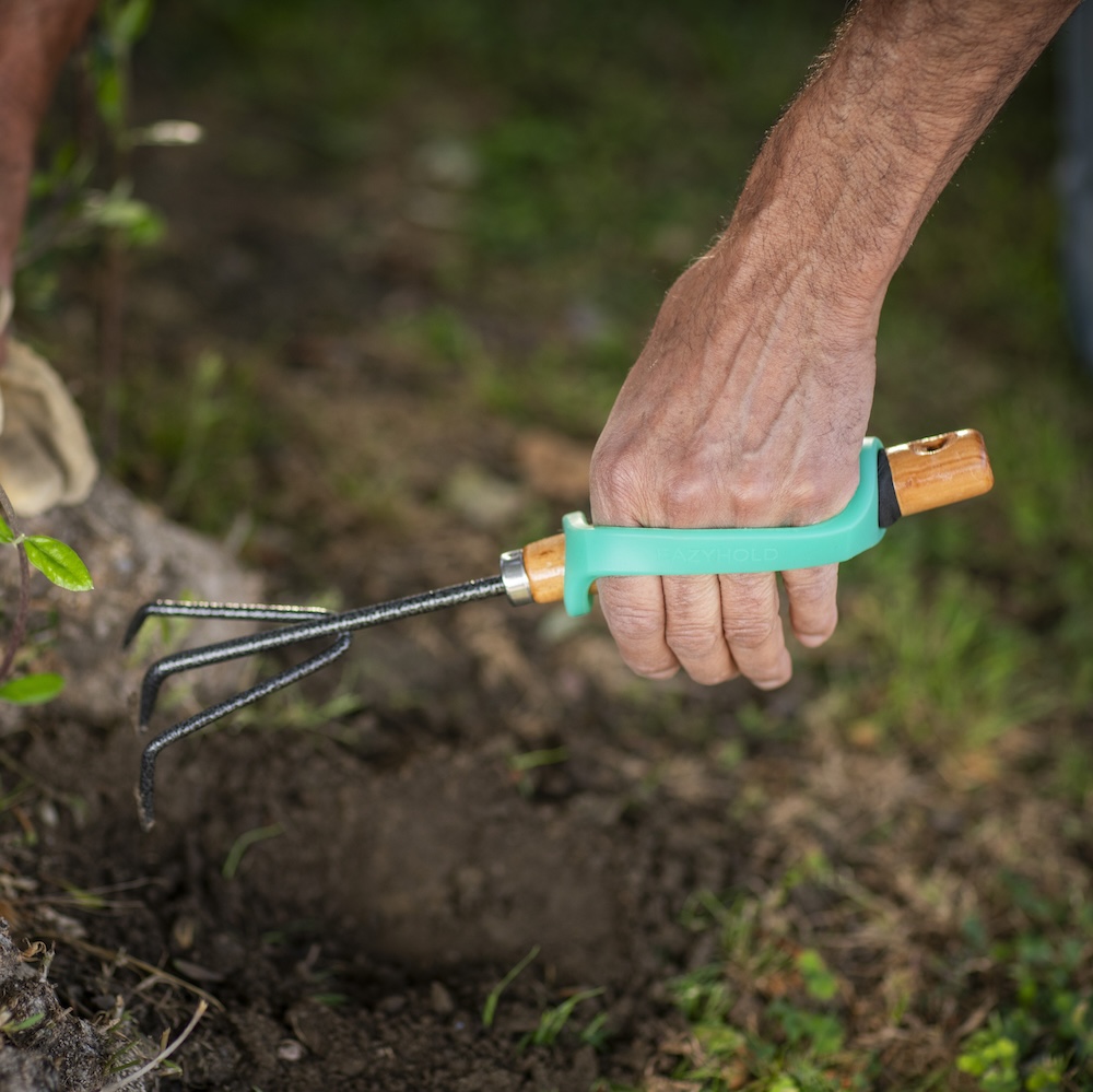 A man's hand uses a small rake over soil with an aqua EasyHold cuff over the back of the hand.