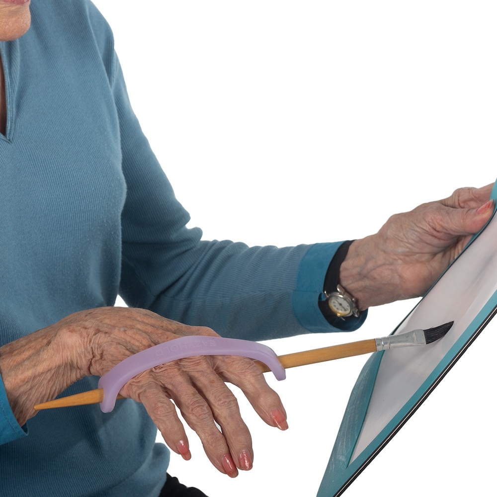 An elderly woman on a white background paints on paper using a lavender EasyHold strap to held her hold her paintbrush.