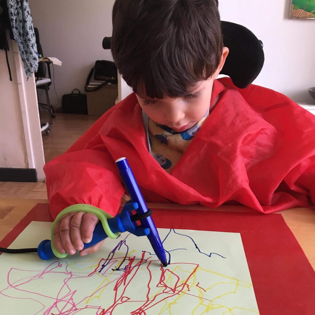 A young boy uses a blue FunctionalHand handle with a green EasyHold cuff on it to hold his marker and draw.