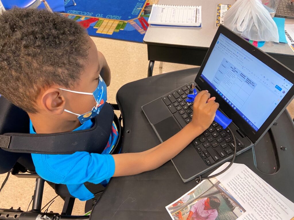 A young boy in school uses a blue FunctionalHand device to hold his stylus and type on a laptop keyboard.