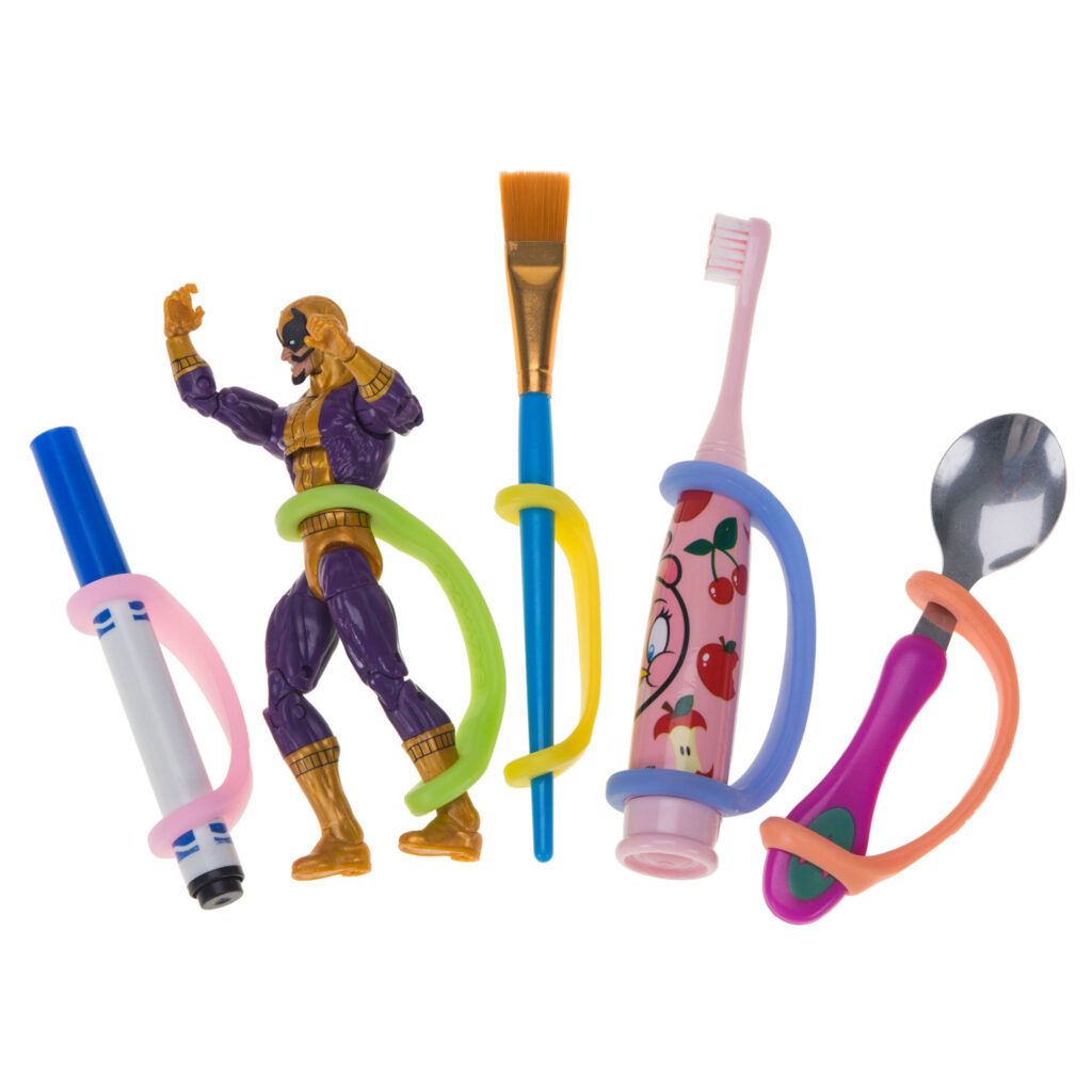 Five silicone universal cuffs in different sizes on toys and tools including a marker, action figure, paintbrush, electric toothbrush, and spoon. The cuffs create a handle and the varying sizes are pink, green, yellow, blue, and orange.