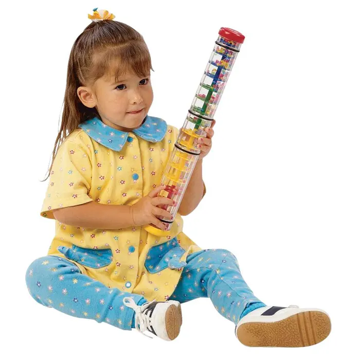 A young girl sits over a white background and holds a long rainbow colored rainstick.