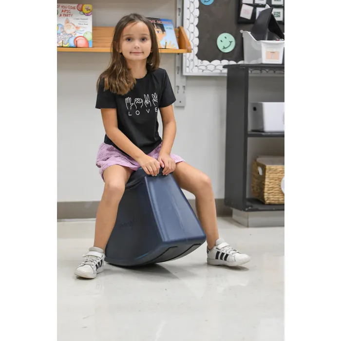 A young girl in a classroom sits straddling a rocking wedge shaped saddle stool.