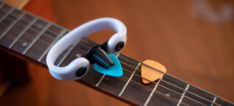 A grey and black 3D printed pick-holding device with an EasyHold universal cuff attached sits atop the neck of a guitar.
