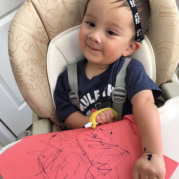 A small boy sits in a supportive chair and draws on red construction paper with a marker equipped with a yellow EasyHold universal cuff.