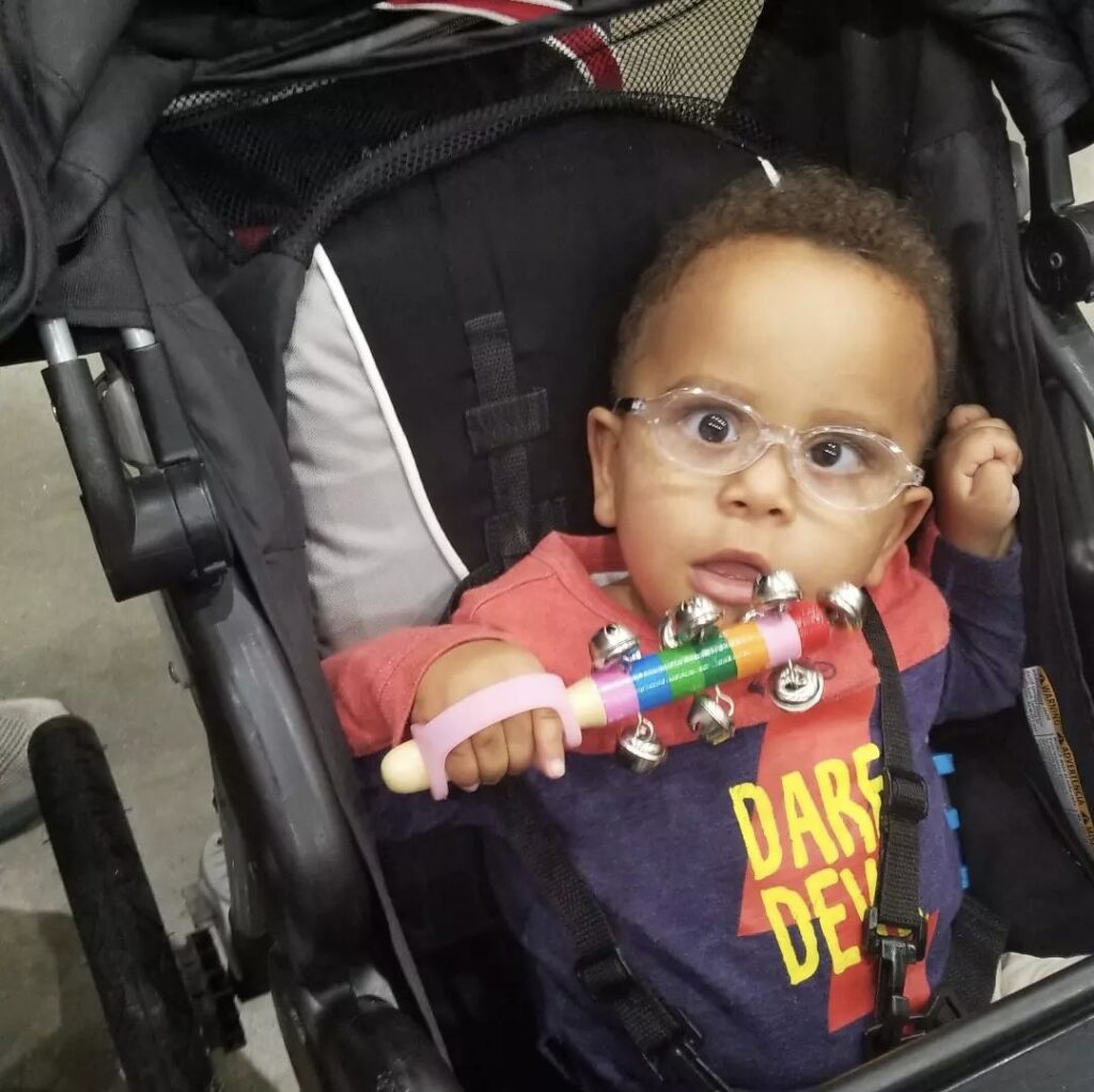 A toddler boy in a stroller holds a set of handled sleigh bells with a small pink EasyHold universal cuff hugging the back of his hand and helping him grasp.